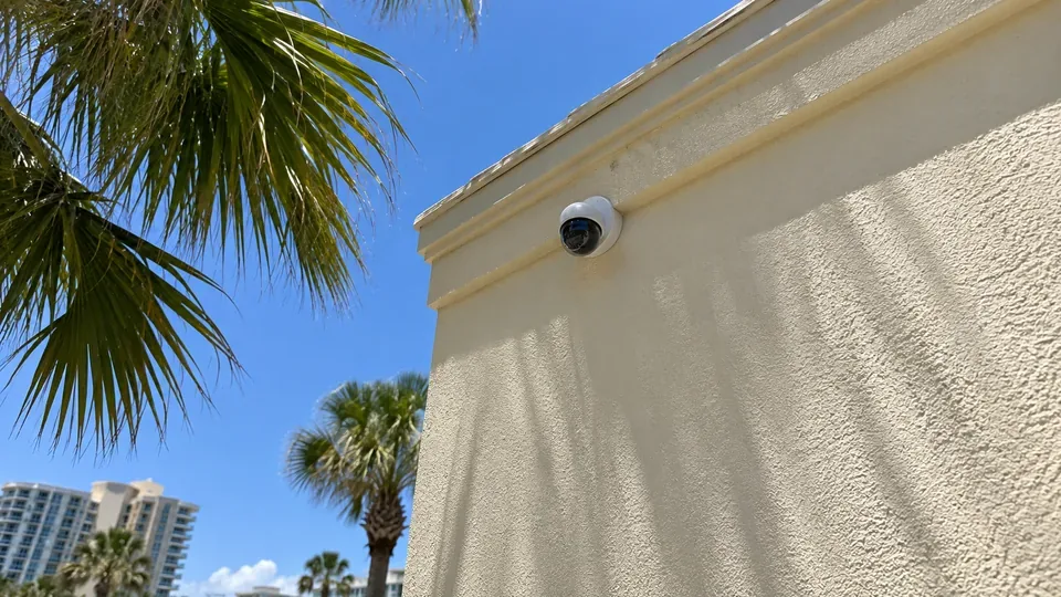 White IP dome camera mounted flush on a stucco commercial wall with palm fronds and blue sky in Daytona Beach