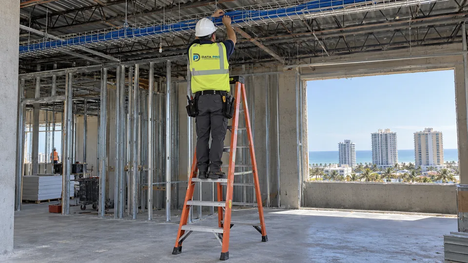Data Pro technician in hard hat and vest on an orange ladder laying low-voltage cable into an overhead basket tray on a coastal commercial build-out